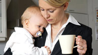Eine Frau im Hosenanzug hält in einer Hand eine Tasse Kaffee, in der anderen ein Baby